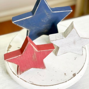 Puede incluir: Tres estrellas de madera pintadas en rojo, blanco y azul, sentadas sobre una base de madera blanca. Las estrellas son una decoraci&oacute;n patri&oacute;tica para el hogar.