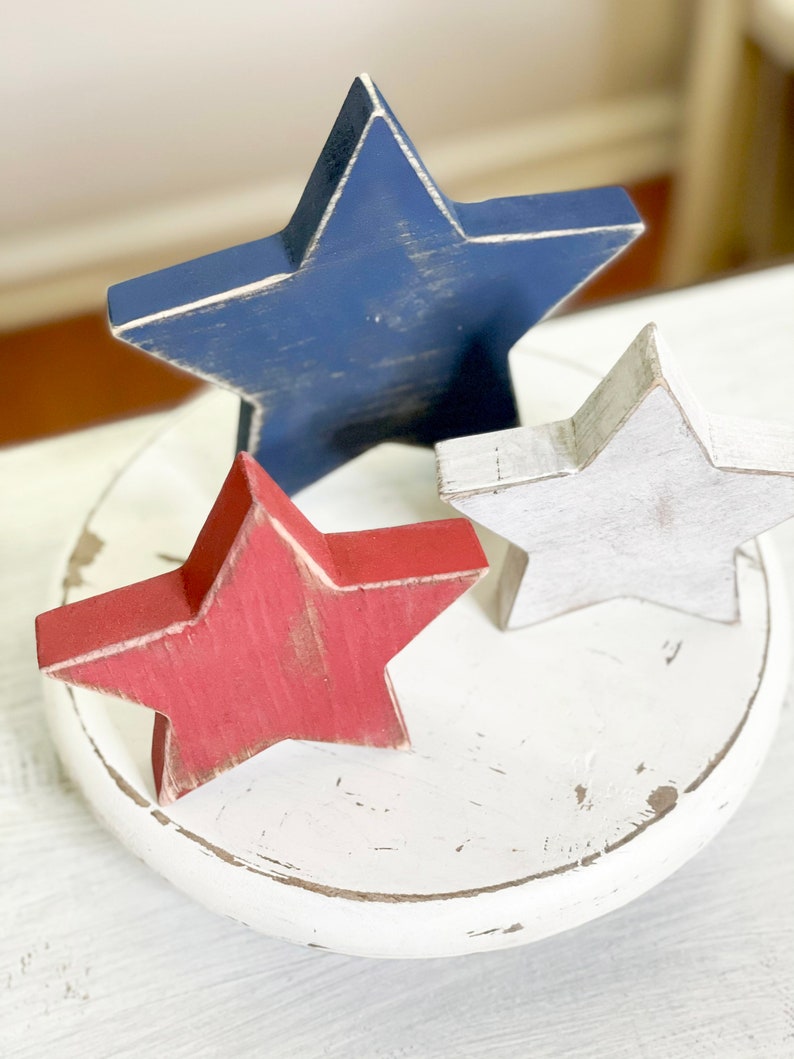Puede incluir: Tres estrellas de madera pintadas en rojo, blanco y azul, sentadas sobre una base de madera blanca. Las estrellas son una decoraci&oacute;n patri&oacute;tica para el hogar.