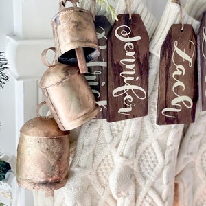 May include: A set of gold-colored bells with wooden tags hanging from a white knitted stocking. The tags are personalized with the names "Jennifer" and "Laci".
