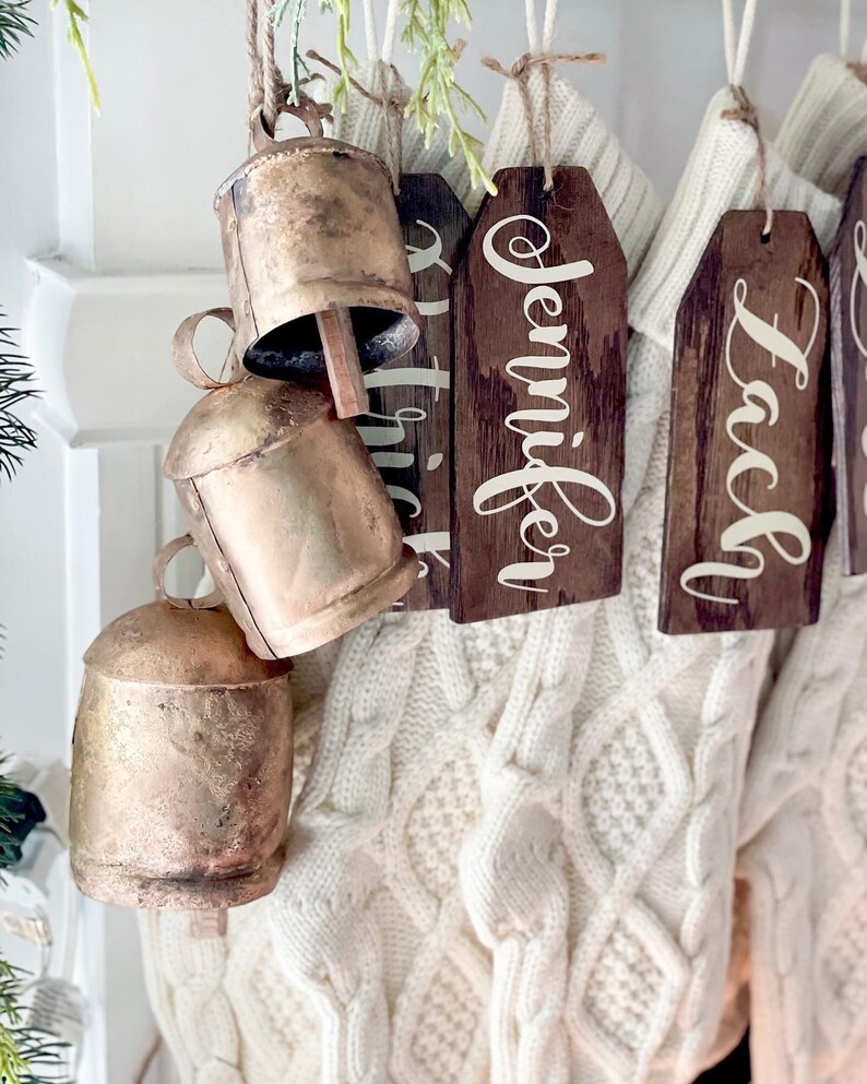 May include: A set of gold-colored bells with wooden tags hanging from a white knitted stocking. The tags are personalized with the names "Jennifer" and "Laci".