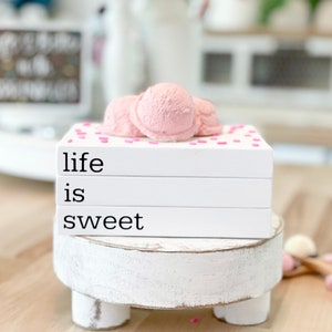 May include: A white wooden tiered stand with three legs, featuring a stack of three white books with black text that reads "life is sweet". The books are topped with three pink ice cream scoops and pink sprinkles.