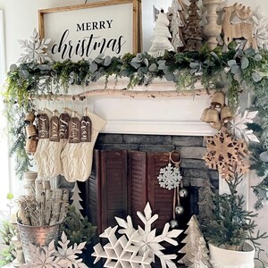 May include: A white wooden sign with the text "Merry Christmas" in black lettering, decorated with a garland of greenery and white snowflakes. The sign is hanging above a fireplace mantel with stockings and other Christmas decorations.