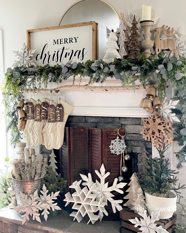 May include: A white wooden sign with the text "Merry Christmas" in black lettering, decorated with a garland of greenery and white snowflakes. The sign is hanging above a fireplace mantel with stockings and other Christmas decorations.