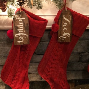 May include: Two red knitted Christmas stockings with wooden name tags. The stockings are hanging from a fireplace mantel. The name tags read "Jennifer" and "Lacy".