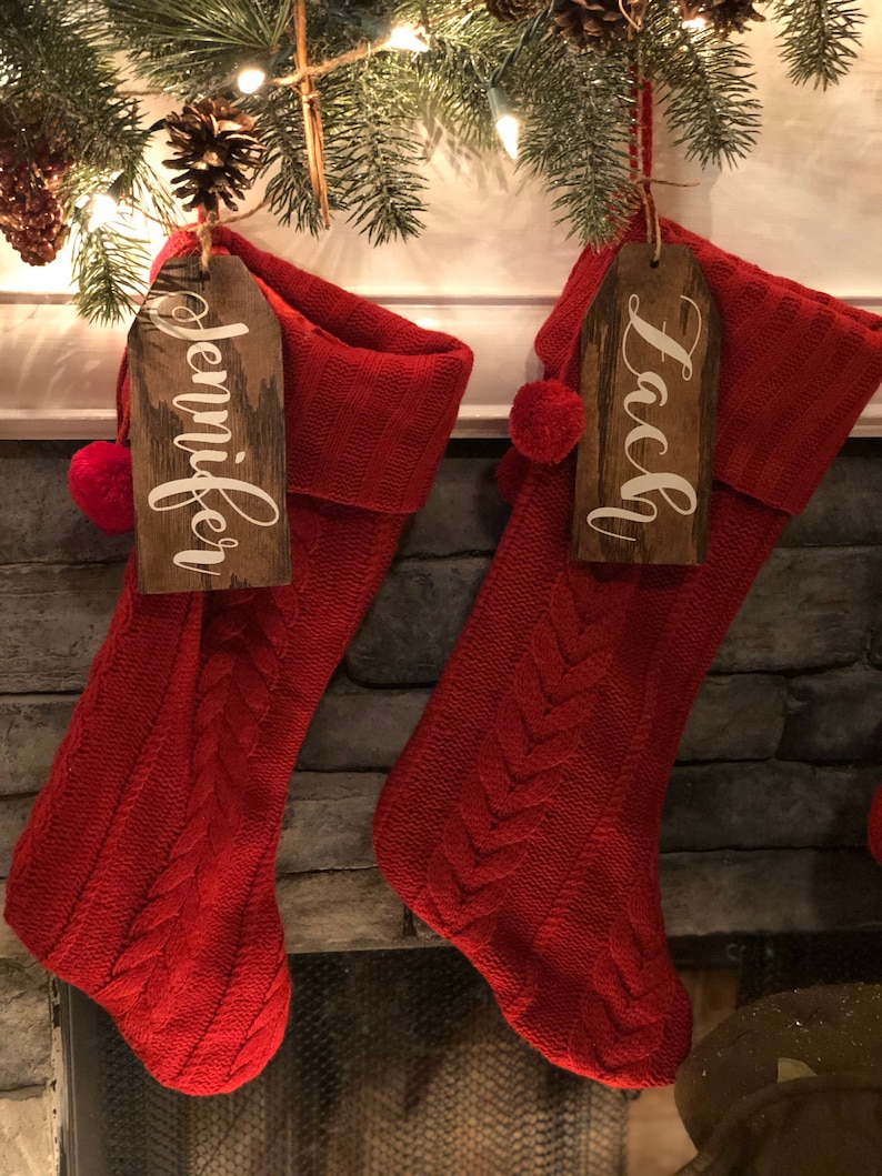May include: Two red knitted Christmas stockings with wooden name tags. The stockings are hanging from a fireplace mantel. The name tags read "Jennifer" and "Lacy".