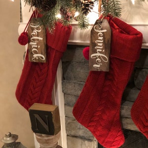 May include: Two red knitted Christmas stockings with wooden name tags that read "Jennifer" and "Patrick". The stockings are hanging on a brick fireplace mantel.