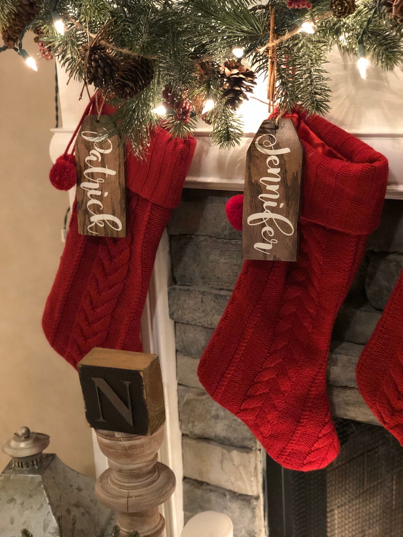 May include: Two red knitted Christmas stockings with wooden name tags that read "Jennifer" and "Patrick". The stockings are hanging on a brick fireplace mantel.
