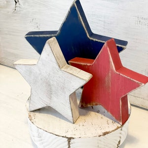 Puede incluir: Tres estrellas de madera en colores rojo, blanco y azul, desgastadas y pintadas, sobre un pedestal de madera blanco.