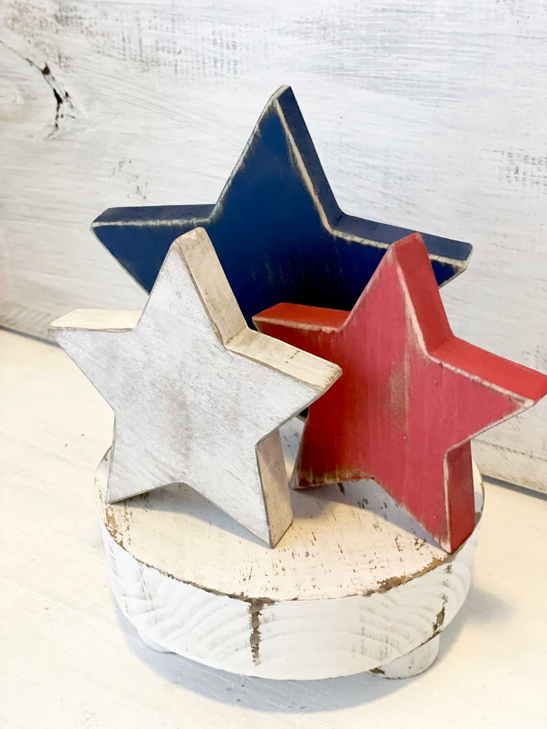 Puede incluir: Tres estrellas de madera en colores rojo, blanco y azul, desgastadas y pintadas, sobre un pedestal de madera blanco.