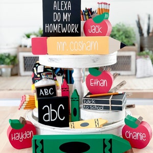 May include: A tiered wooden display featuring school-themed decorations. Includes a black sign with "ALEXA DO MY HOMEWORK", a pencil-shaped sign with "mr. cosham", and red apple-shaped signs with names. Other items include crayons and a sign that says "back to school".