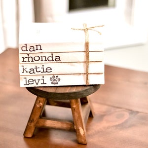 May include: A stack of four white books with the names "dan", "rhonda", "katie", and "levi" printed on them. The bottom book has a paw print next to the name "levi". The books are tied together with twine and are sitting on a small wooden stool.