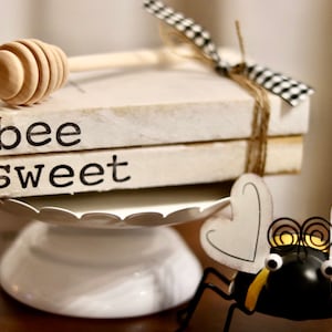 May include: Two white books stacked on a white pedestal with the words "bee sweet" printed on the front. A wooden honey dipper is resting on top of the books. A black and yellow bee figurine with white wings is in the foreground.