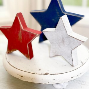 Puede incluir: Tres estrellas de madera en rojo, blanco y azul, sentadas sobre un pedestal blanco. Las estrellas est&aacute;n pintadas con un acabado desgastado.