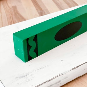 May include: A green wooden crayon-shaped block with a black oval and black squiggly lines. The block sits on a white, distressed wooden surface. The background shows a wooden floor.