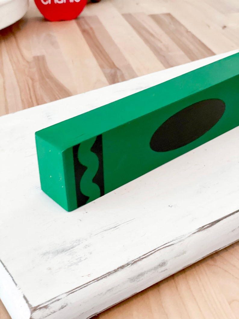 May include: A green wooden crayon-shaped block with a black oval and black squiggly lines. The block sits on a white, distressed wooden surface. The background shows a wooden floor.