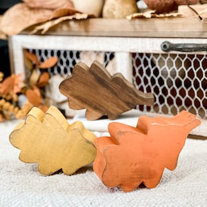 May include: Three wooden oak leaf shapes painted in different fall colors: brown, yellow, and orange. The leaves are arranged on a white textured surface.