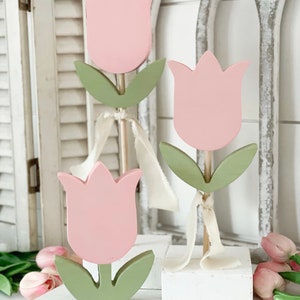 May include: Three wooden tulip decorations. Each tulip has a pink flower head, green leaves, and a wooden stem. Cream-colored ribbon is tied around the stems. The tulips are displayed on a white surface with a white background.