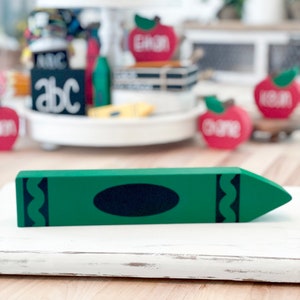 May include: A green wooden crayon decoration with a black oval and wavy lines on the sides. The crayon is placed on a white surface. Other school-themed decorations are visible in the background.