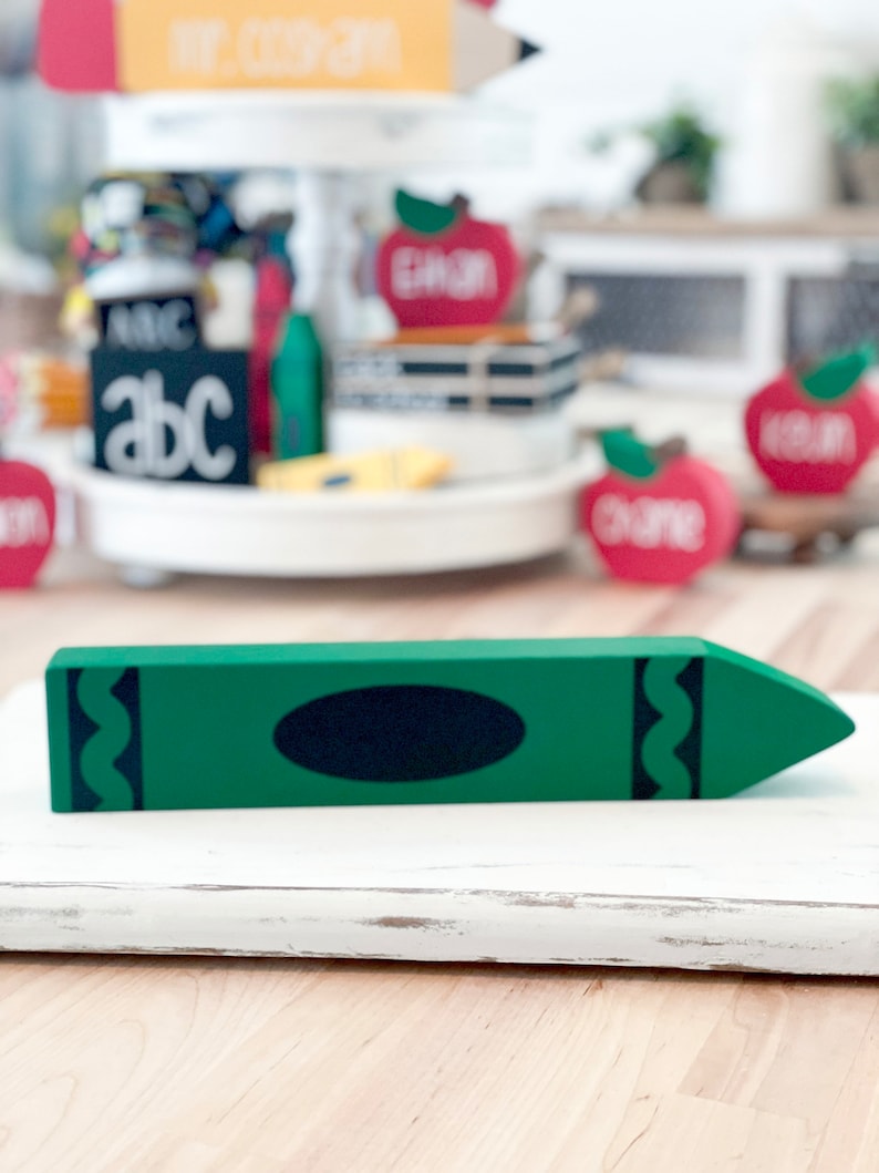 May include: A green wooden crayon decoration with a black oval and wavy lines on the sides. The crayon is placed on a white surface. Other school-themed decorations are visible in the background.