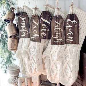 May include: Five wooden Christmas stocking tags with names written in white script. The names are Jennifer, Jack, Lovie, Taliegh, and Evan. The tags are hanging from white twine on a white knitted stocking.