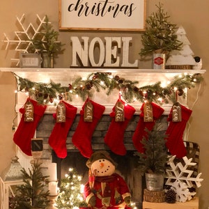 May include: A white fireplace mantel decorated for Christmas with a sign that reads "Merry Christmas" and "Noel" in large white letters. There are stockings hung on the mantel with names written on them. A snowman figurine sits in front of the fireplace with a small Christmas tree and lights.