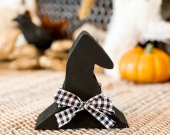 May include: Black wooden witch hat with a black and white checkered ribbon bow. The hat is sitting on a woven rug.