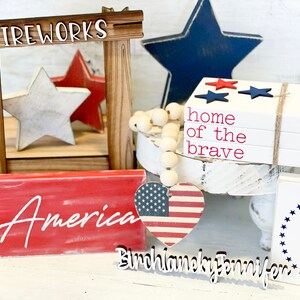 Puede incluir: Una decoraci&oacute;n para el hogar con tem&aacute;tica patri&oacute;tica con un letrero de madera que dice "FIREWORKS", una estrella roja y blanca, una estrella azul, una pila de bloques de madera blanca con las palabras "home of the brave" en rojo, un bloque de madera rojo con la palabra "America" en blanco, un coraz&oacute;n rojo, blanco y azul con un letrero de madera que dice "Birchlaneky Jennifer", y un bloque de madera blanco con las palabras "God Bless America" en rojo y azul.