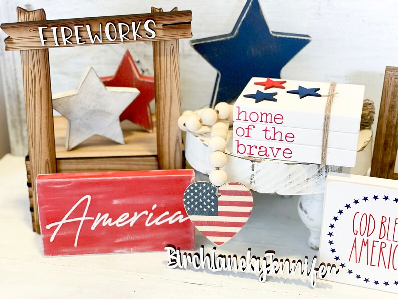 Puede incluir: Una decoraci&oacute;n para el hogar con tem&aacute;tica patri&oacute;tica con un letrero de madera que dice "FIREWORKS", una estrella roja y blanca, una estrella azul, una pila de bloques de madera blanca con las palabras "home of the brave" en rojo, un bloque de madera rojo con la palabra "America" en blanco, un coraz&oacute;n rojo, blanco y azul con un letrero de madera que dice "Birchlaneky Jennifer", y un bloque de madera blanco con las palabras "God Bless America" en rojo y azul.