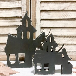 May include: Two black wooden haunted house decorations with white distressed edges. The houses have cut-out windows and doors, and are placed on a white surface in front of a white shutter background. The houses are a Halloween or spooky themed decoration.