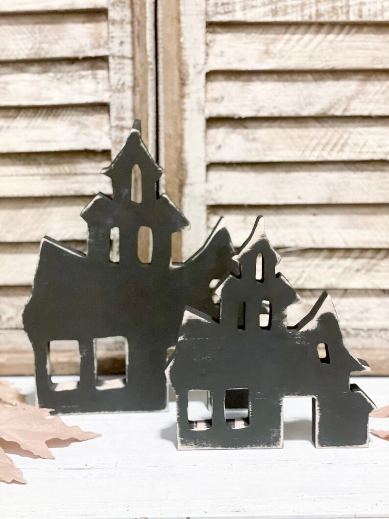 May include: Two black wooden haunted house decorations with white distressed edges. The houses have cut-out windows and doors, and are placed on a white surface in front of a white shutter background. The houses are a Halloween or spooky themed decoration.