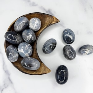 May include: A collection of oval-shaped blue-grey stones with white patterns, displayed in a wooden crescent-shaped bowl. Some stones are scattered on a white marble surface. The stones have a polished appearance, suitable for decorative purposes.