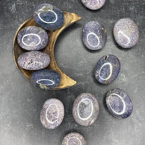 May include: A collection of polished, oval-shaped stones in shades of purple and gray. Some stones are displayed in a wooden crescent-shaped bowl. The stones have a smooth, glossy finish and varying patterns. The background is a dark gray surface.
