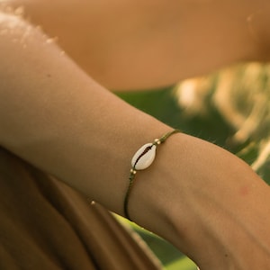 Può includere: Un braccialetto di corda verde con un ciondolo a forma di conchiglia di cauri bianca.