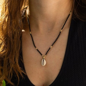 Könnte beinhalten: Eine schwarze Perlenkette mit goldenen Akzenten und einem weißen Muschel-Anhänger. Die Halskette wird um den Hals getragen und ist ein schlichtes, elegantes Schmuckstück.