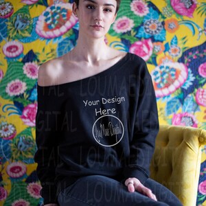 Può includere: Felpa nera con spalle scoperte con la scritta "Your Design Here" e un logo circolare. La persona è seduta su una sedia gialla contro uno sfondo a motivi floreali. La felpa è abbinata a jeans scuri.