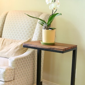 May include: A black metal side table with a wooden top. The table has a simple, modern design and is perfect for a living room or bedroom. A potted white orchid sits on the table.