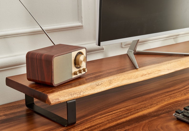 May include: A wooden monitor stand with a black metal frame. A vintage-style radio with a wooden case and gold accents sits on the stand. The stand is on a wooden desk.