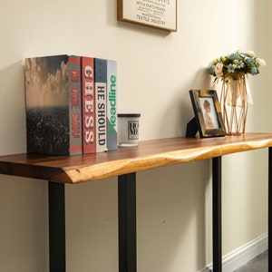 May include: A wooden console table with a live edge top and black metal legs. The table is decorated with a vase of flowers, a framed photo, and a stack of books with titles "The Shop", "He Should", "Chess", "My Time", and "Headline".