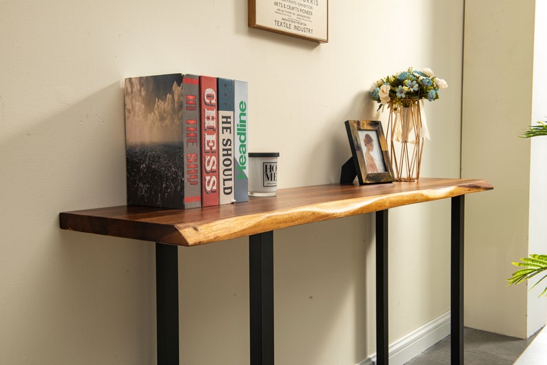 May include: A wooden console table with a live edge top and black metal legs. The table is decorated with a vase of flowers, a framed photo, and a stack of books with titles "The Shop", "He Should", "Chess", "My Time", and "Headline".