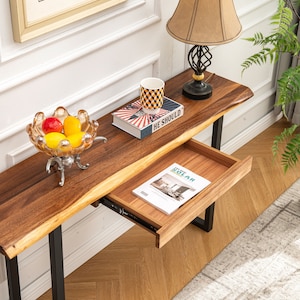 May include: A wooden console table with a live edge top and black metal legs. The table has a drawer that is open and a book is inside. There is a bowl of fruit on the table and a lamp with a beige shade on the table.