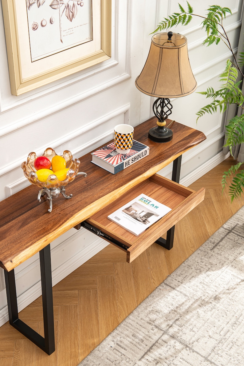May include: A wooden console table with a live edge top and black metal legs. The table has a drawer that is open and a book is inside. There is a bowl of fruit on the table and a lamp with a beige shade on the table.