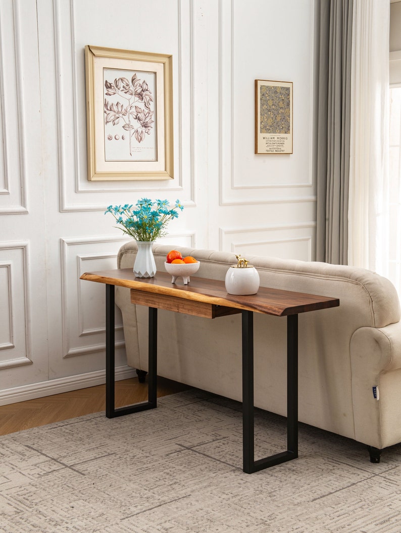 May include: A wooden console table with a live edge top and black metal legs. The table has a drawer and is sitting on a patterned rug in front of a white sofa.