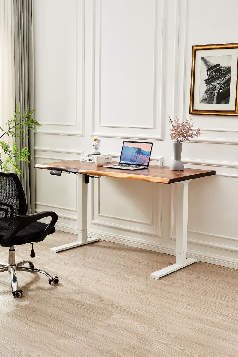 May include: A white standing desk with a wooden top and a black office chair. The desk has a white frame and a white base. The desk is in a room with white walls and a wooden floor. There is a laptop computer, a vase with flowers, and a book on the desk.