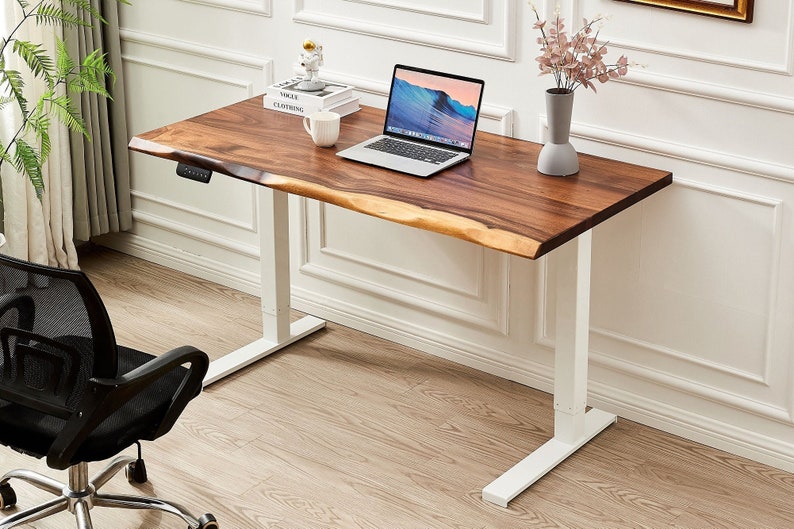May include: A white standing desk with a wooden top. The desk has a natural edge and a white metal frame. A laptop computer, a mug, and a book are on the desk.