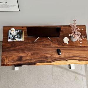 May include: A dark wood desk with a live edge top and white metal legs. A computer monitor, a cup, a mouse, and a vase with pink flowers are on the desk.