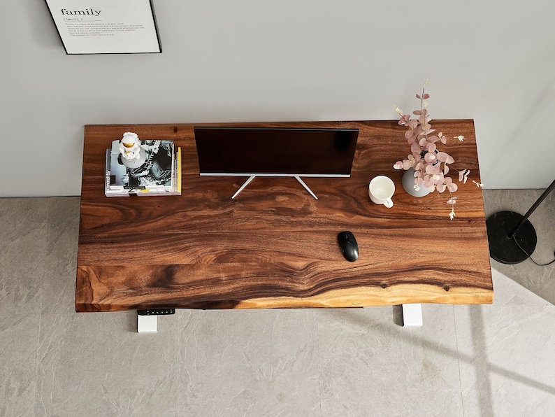 May include: A dark wood desk with a live edge top and white metal legs. A computer monitor, a cup, a mouse, and a vase with pink flowers are on the desk.