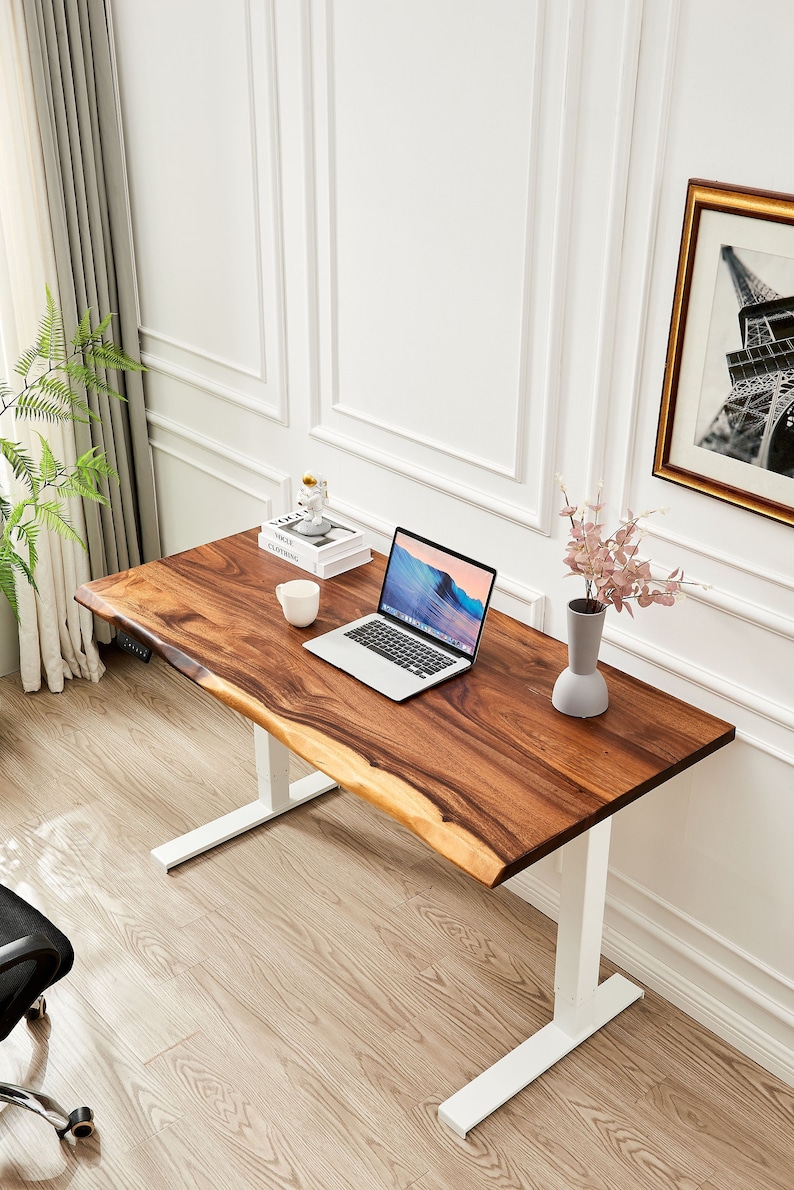 May include: A white standing desk with a wooden top. The desk has a laptop, a mug, a small sculpture, and a vase of flowers on it. The desk is in a room with white walls and a wooden floor.