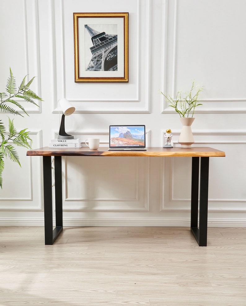 Pode incluir: Uma mesa de madeira com pernas de metal preto. A mesa tem um laptop, uma x&iacute;cara de caf&eacute;, um livro e uma l&acirc;mpada. H&aacute; uma imagem emoldurada da Torre Eiffel na parede atr&aacute;s da mesa.