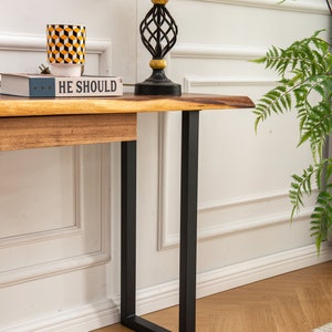 May include: A wooden console table with a live edge top and black metal legs. The table has a book and a decorative cup on top. The table is in front of a white wall with white trim.