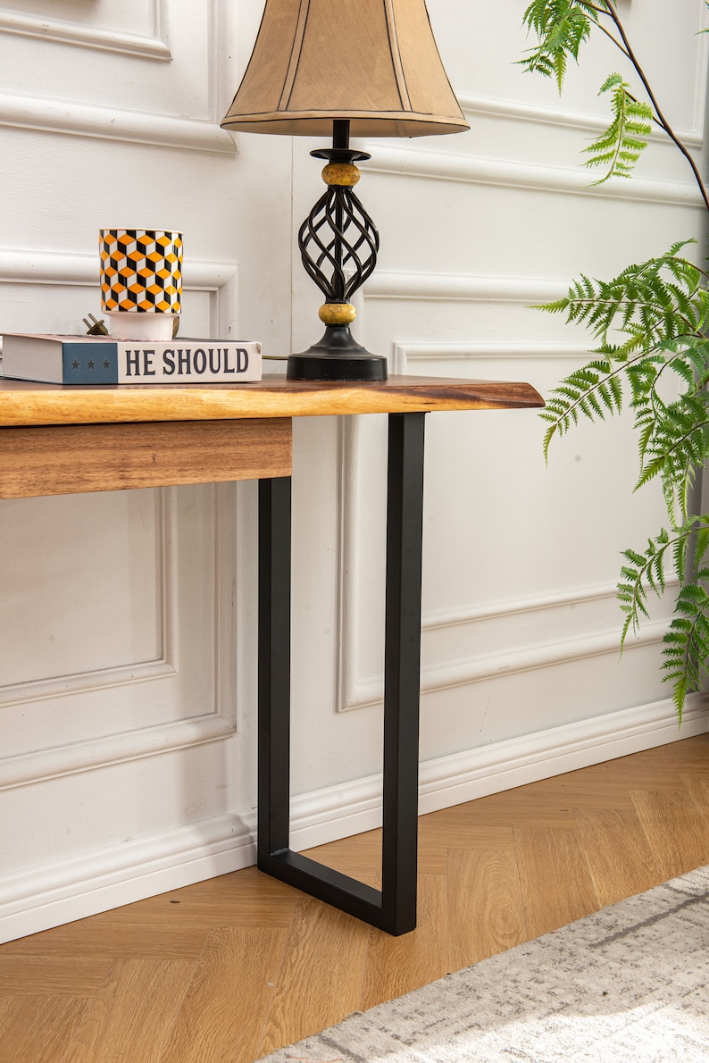 May include: A wooden console table with a live edge top and black metal legs. The table has a book and a decorative cup on top. The table is in front of a white wall with white trim.